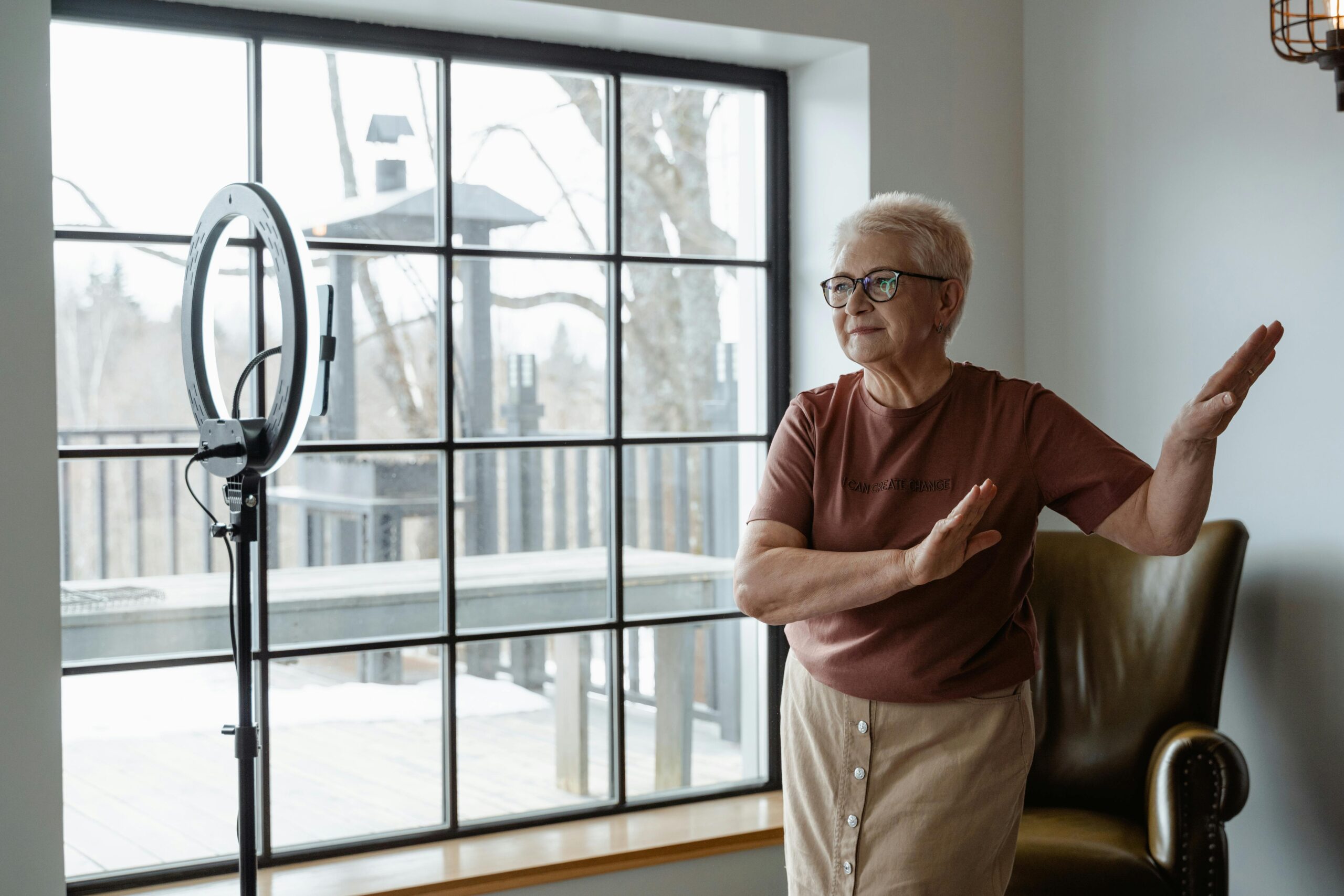 Elderly woman joyfully dancing indoors near a large window with ring light.