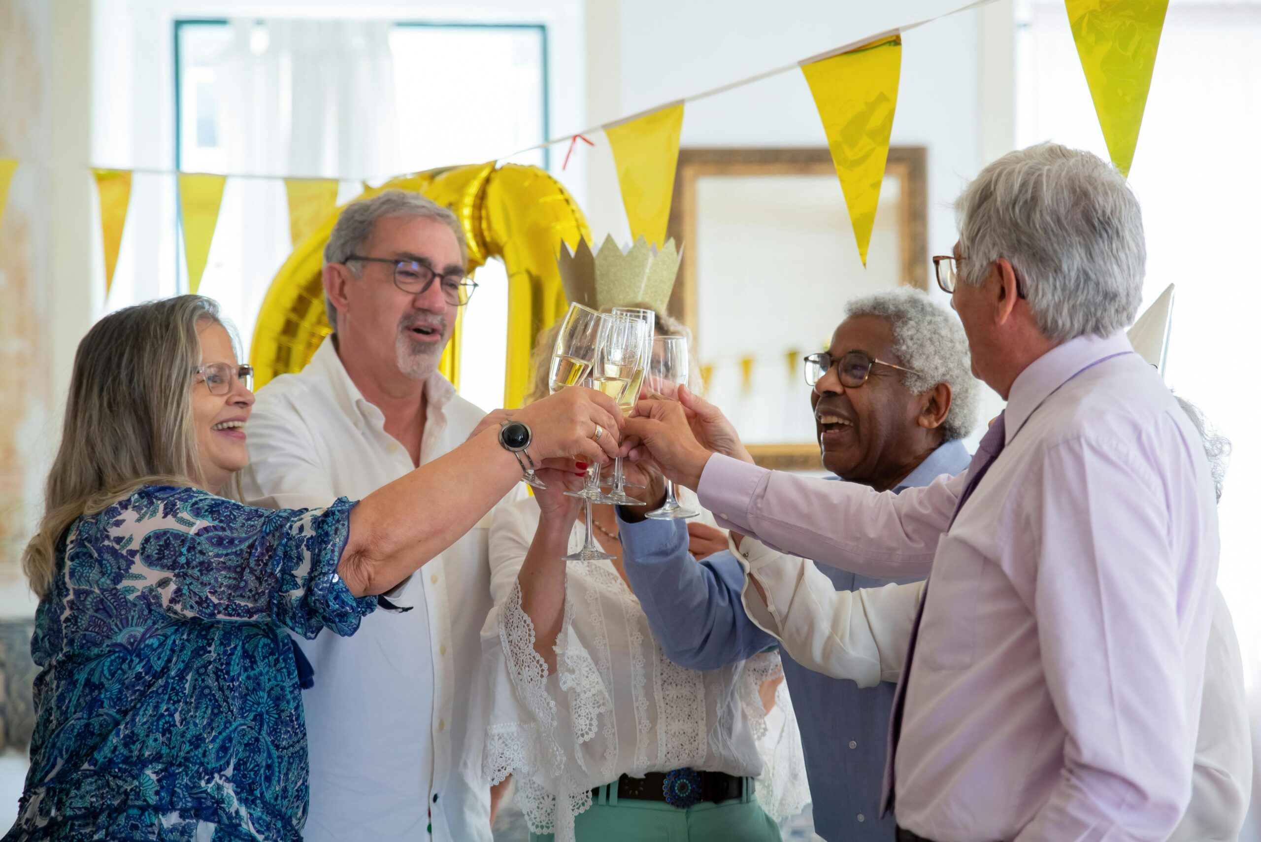 A group of diverse seniors celebrating indoors, toasting with champagne flutes under festive yellow decor.